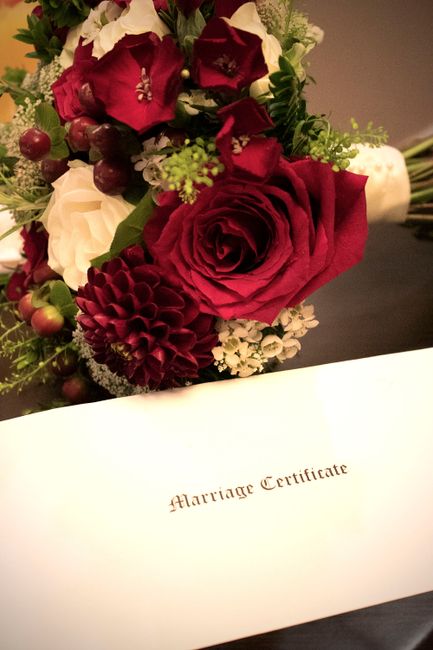 A close-up of a floral arrangement featuring a large, deep red rose at the center, surrounded by burgundy and white flowers including roses and dahlias, with green foliage and small red berries. The b