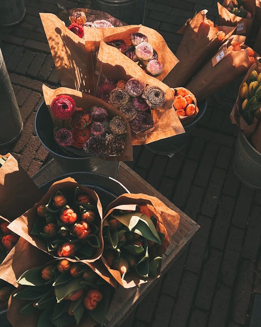 An outdoor display of fresh flower bouquets arranged with variety and care, featuring vibrant colors such as deep reds, purples, and oranges. The bouquets are wrapped in rustic brown paper and are pla