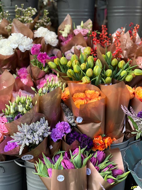 A vibrant display of freshly cut flowers arranged in brown paper wrappings, featuring a variety of blooms including pink and white carnations, purple statice, green tulip buds, orange roses, and laven